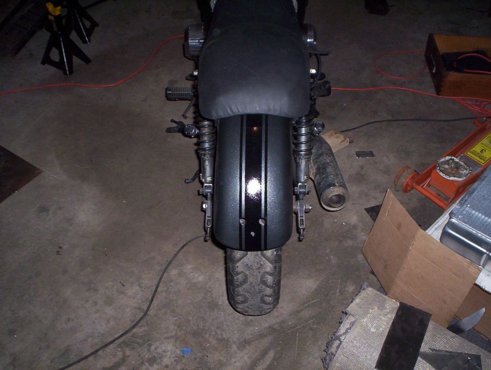 Motorcycle rear fender with black and silver stripes, showing the tire and part of the exhaust pipe. A floor jack is visible to the right