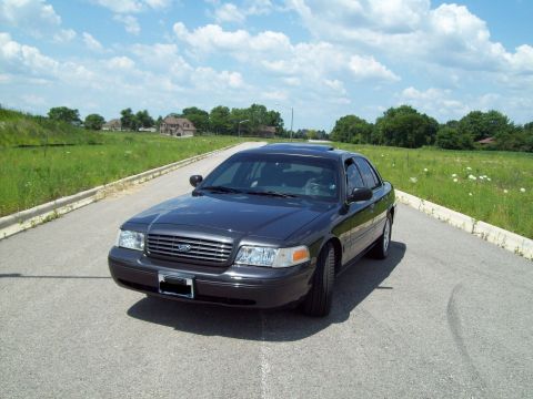 2004 Ford Crown Victoria - Dark gray sedan with a sun visor on the windshield sits on an asphalt road with a grassy shoulder.