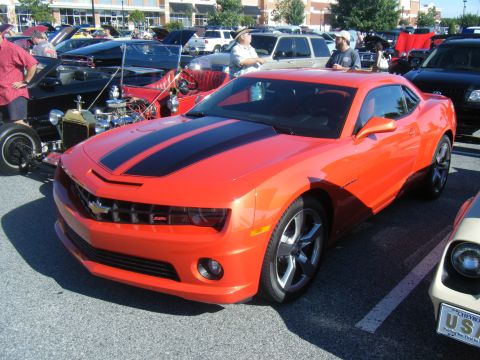 1991 Nissan Micra - An orange Chevrolet Camaro with black racing stripes is parked at a car show, with other vehicles visible in the background.