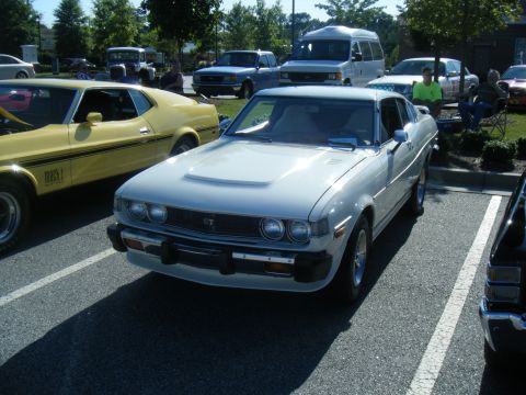 1991 Nissan Micra - A white 1975 Toyota Celica GT coupe is parked in a lot next to a yellow Ford Mustang Mach 1.