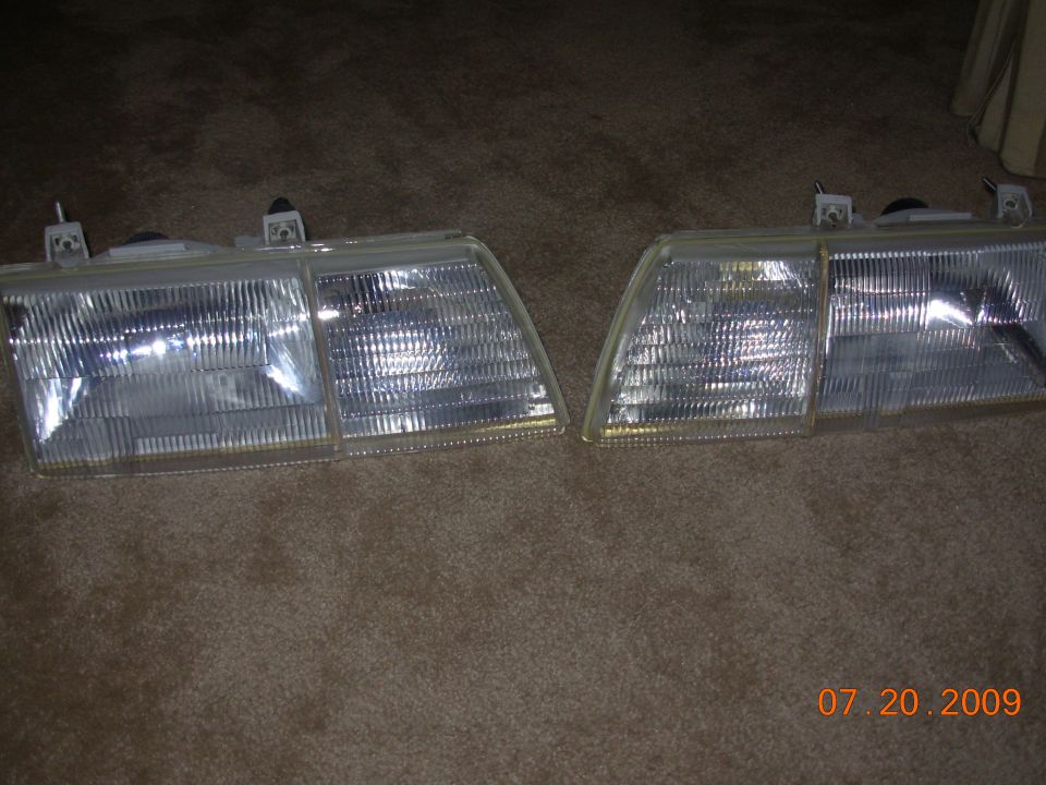 Two clear plastic car headlights with amber turn signal sections, lying on a carpeted surface