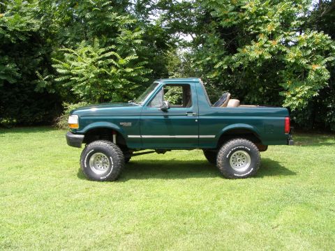 1993 Ford Bronco - Side view of a lifted green 1993 Ford Bronco parked on a grassy lawn with trees in the background.