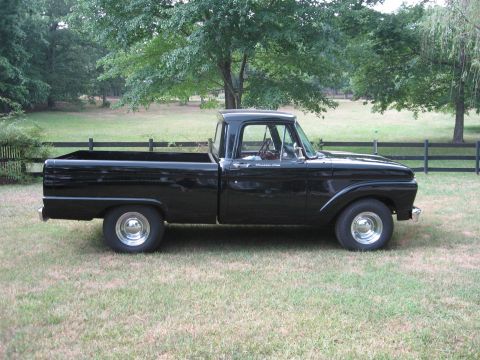 1966 Ford F-100 - A classic black pickup truck is shown from the side, parked on a grassy lawn with trees and a fence behind it.