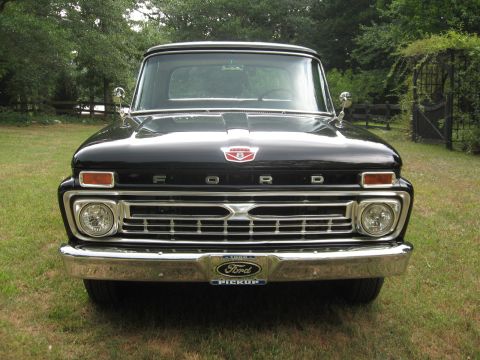 1966 Ford F-100 - A black 1966 Ford F-100 pickup truck is shown from the front, parked on a grassy area with trees in the background.