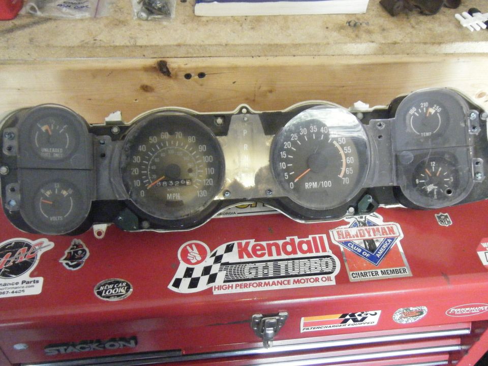 Car dashboard instrument cluster with speedometer, tachometer, fuel gauge, and voltmeter. Stickers on the red toolbox below