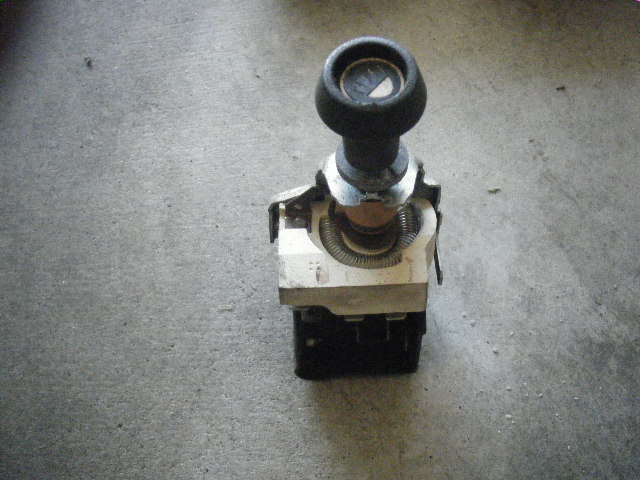 Car part with a black knob on top and a dial visible underneath. It has a metal bracket and a coiled spring