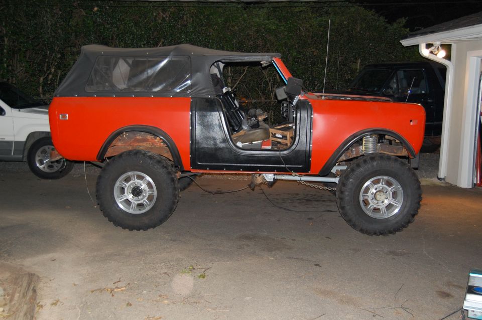 Orange and black International Scout II with a soft top, lifted on large tires. Another SUV is parked to the left