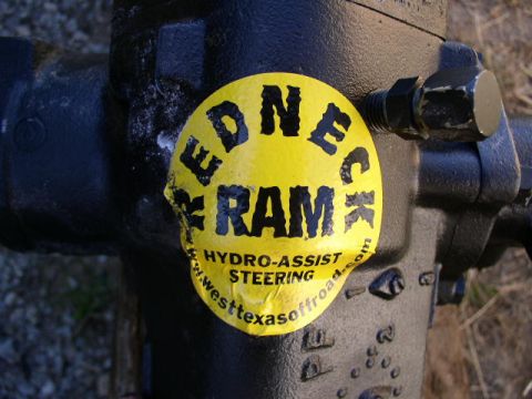 1979 Ford Bronco - Close-up of a steering assist ram with a "Redneck Ram" sticker, indicating hydro-assist steering.