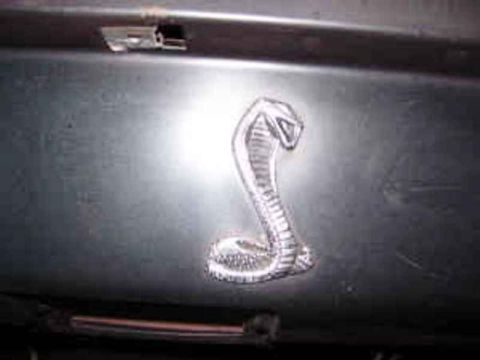 1971 Ford Ranchero - A close-up shows a cobra emblem on a dark, metallic surface, with the Supermotors.net logo in the lower right corner.