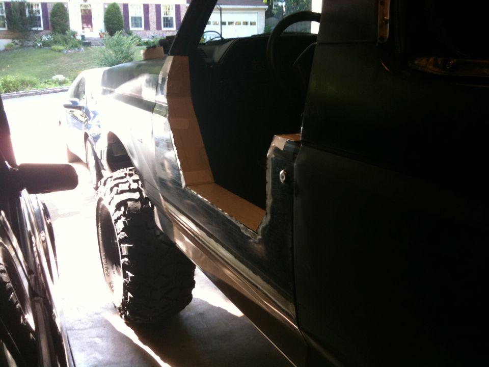 Side view of a dark green Ford Bronco with doors removed, showing interior and large off-road tires. Bodywork is in progress