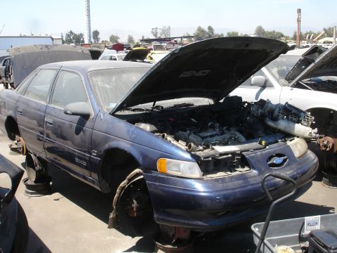 1993 Ford Taurus - A blue 1993 Ford Taurus SHO sits in a junkyard with its hood open and missing wheels.