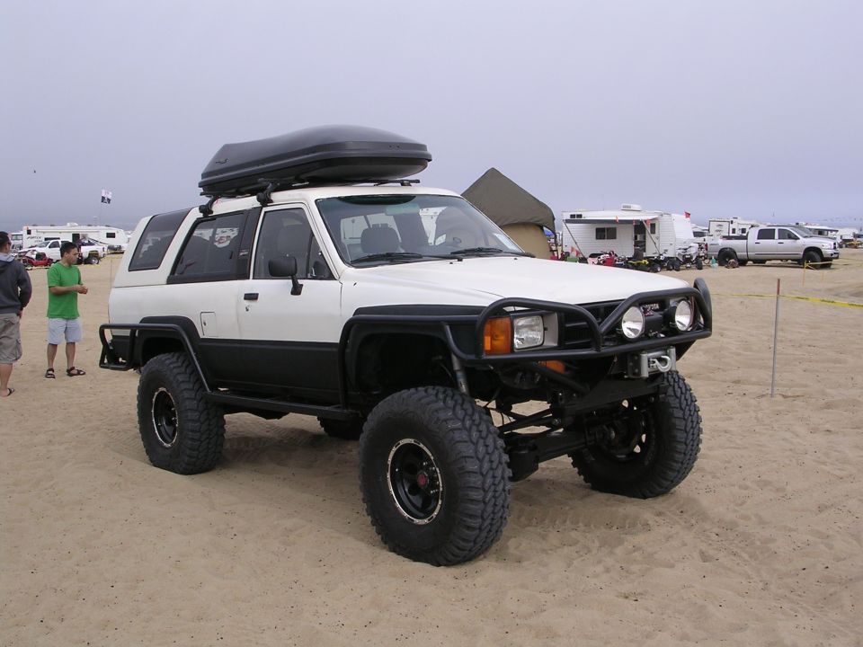 White and black 1985 Toyota 4Runner with a black roof-top cargo carrier, off-road tires, and a front brush guard, parked on sand