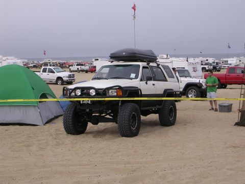 1985 Toyota 4Runner - A modified white 1985 Toyota 4Runner is parked on a beach with a cargo box and large off-road tires, surrounded by other vehicles and tents.