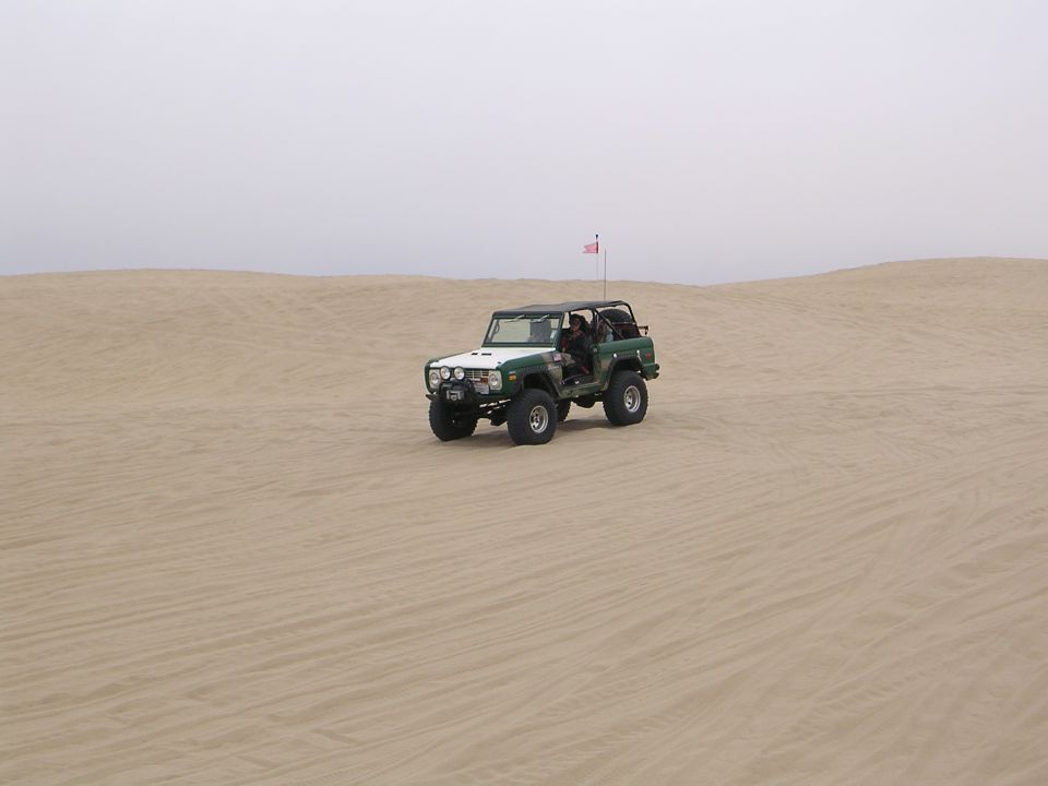 Green and white off-road vehicle with a roll cage driving on sand dunes under a hazy sky