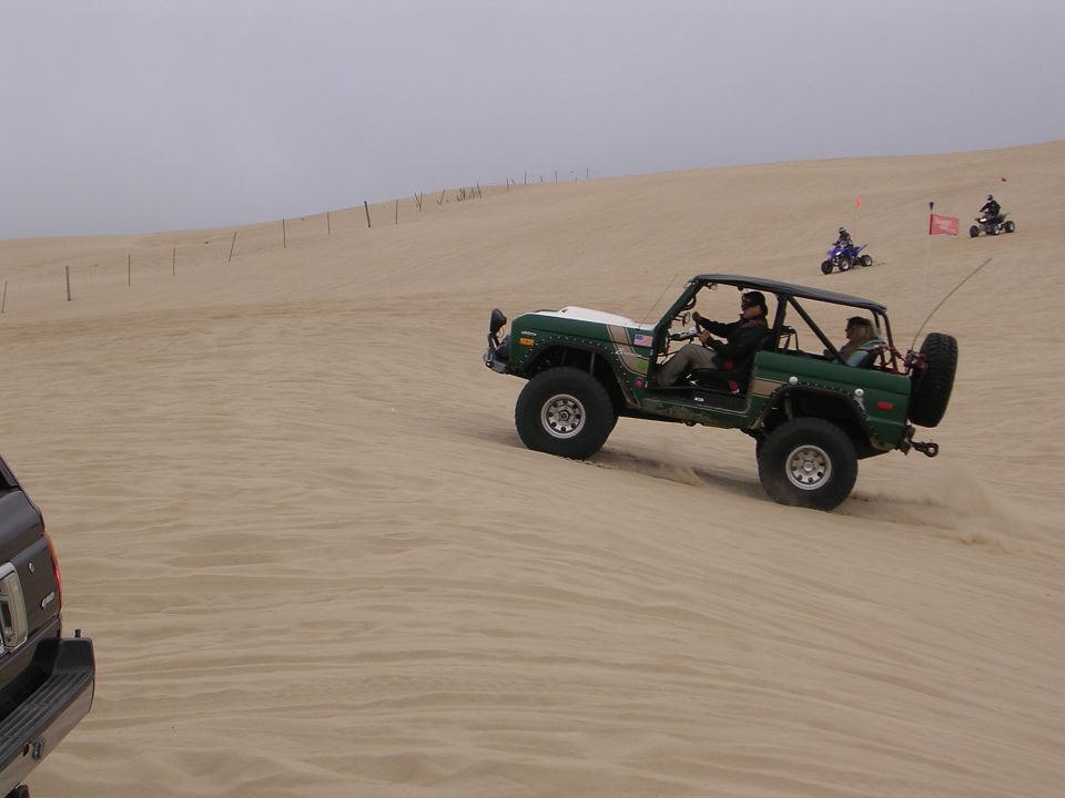 Green off-road vehicle with roll cage driving on sand dunes, with ATVs in the background