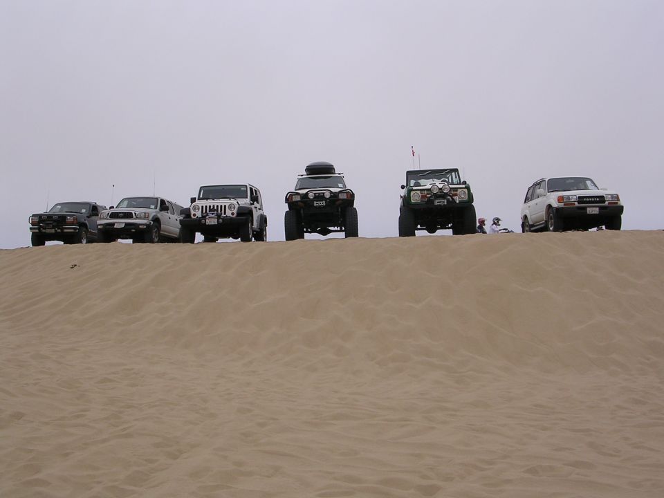 Five off-road vehicles parked on a sand dune under an overcast sky