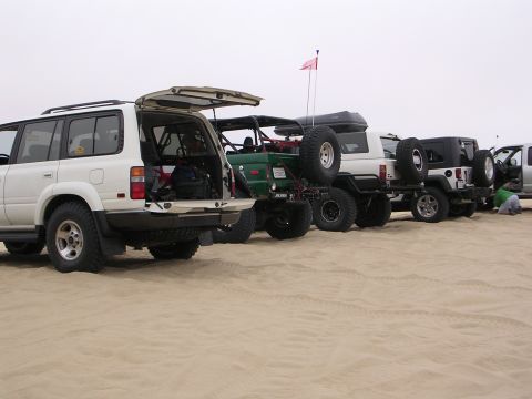 1985 Toyota 4Runner - A line of off-road vehicles, including a white Toyota Land Cruiser, a green custom build, and several Jeeps, are parked on a sandy terrain.