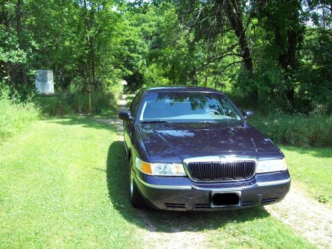 1999 Mercury Grand Marquis - A dark-colored sedan is parked on a grassy area next to a dirt path, surrounded by trees.