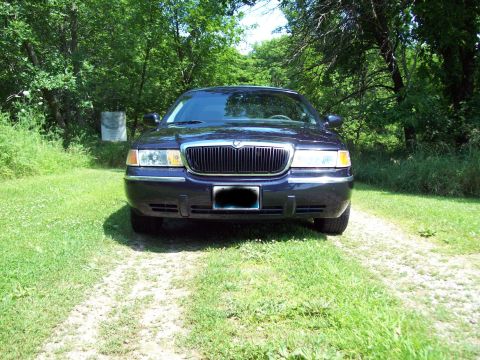 1999 Mercury Grand Marquis - A dark blue sedan is parked on a dirt path surrounded by grass and trees.