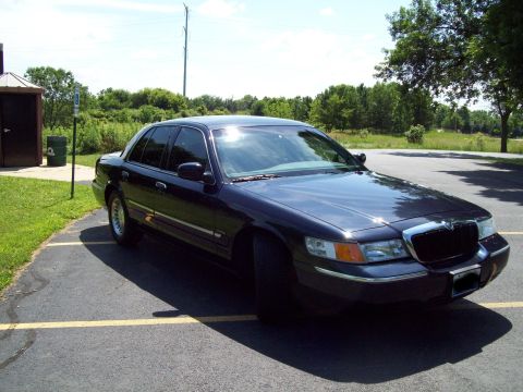 1999 Mercury Grand Marquis - This dark sedan is parked in a lot on a sunny day, with trees and a field in the background.