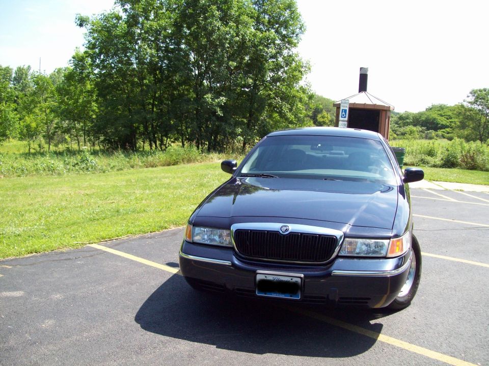 Dark sedan parked in a lot, front view. Chrome grille and trim. Trees and grass in background
