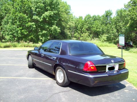 1999 Mercury Grand Marquis - Dark blue Mercury Grand Marquis viewed from the driver-side rear three-quarter angle in an empty asphalt parking lot backed by dense green…