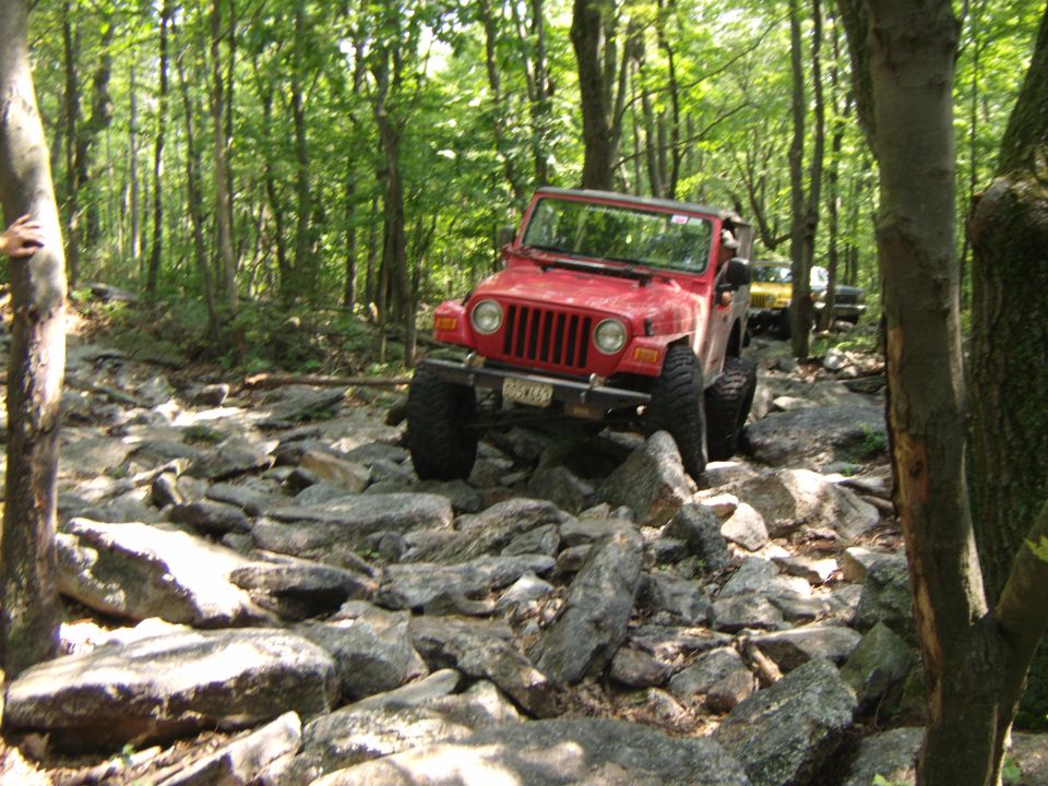 Red 2003 Jeep Wrangler navigating a rocky forest trail with another vehicle behind it