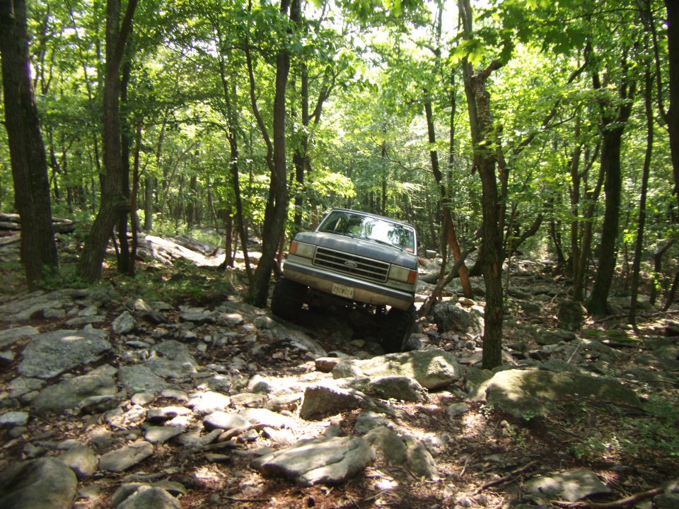 Gray SUV with a prominent grille and large tires navigates a rocky, wooded path