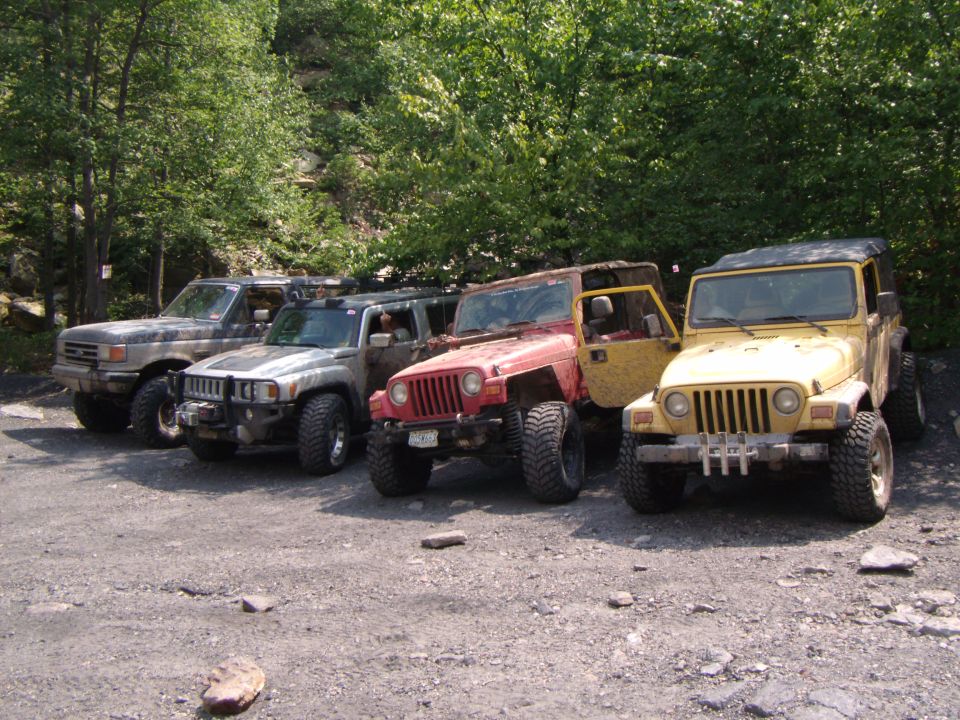 Four modified off-road vehicles parked on gravel: a gray Ford Bronco, a silver Hummer H3, a red Jeep Wrangler TJ, and a yellow Jeep Wrangler TJ
