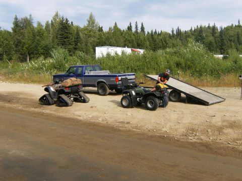 2000 Ford E350 - A pickup truck, ATVs, and a trailer are parked on a dirt road with trees and buildings in the background.