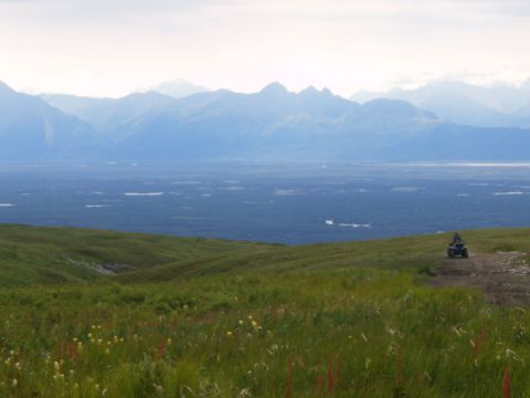 2000 Ford E350 - An ATV sits on a dirt trail, with rolling green hills and distant mountains under a hazy sky.