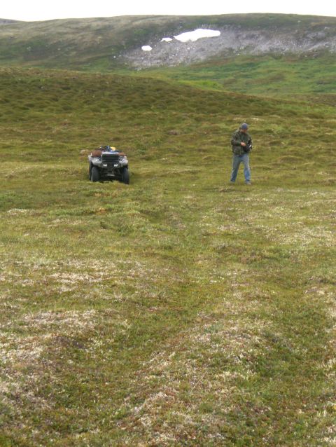 2000 Ford E350 - A person stands in a grassy field next to an ATV, with a snow-patched hill in the background.