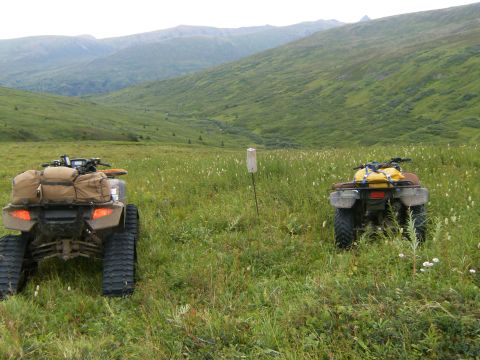 2000 Ford E350 - Two ATVs are parked in a vast, green meadow with a stream visible in the distance and mountains beyond.
