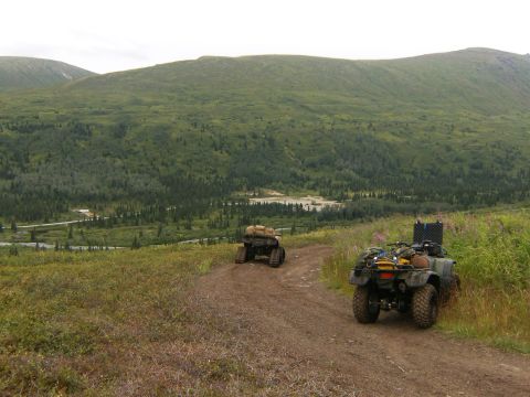 2000 Ford E350 - ATVs traverse a remote dirt road, heading into a vast, green landscape with mountains in the distance.