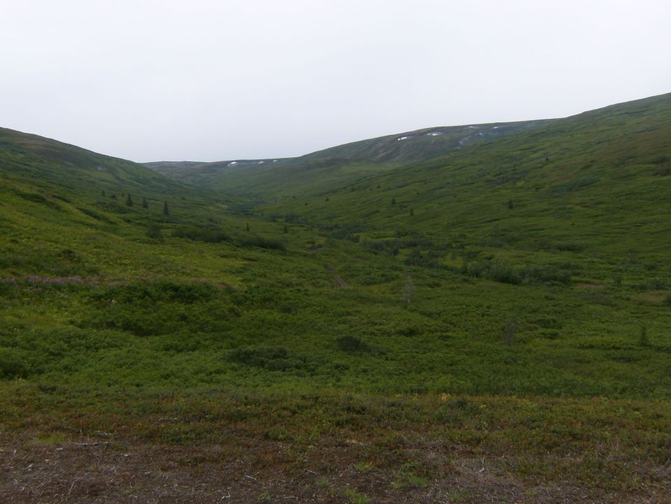 Green hillsides form a valley under a cloudy sky. Some patches of snow are visible on the higher slopes