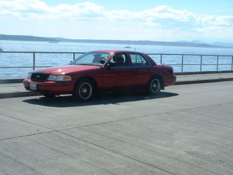 1999 Ford Crown Victoria - A red 1999 Ford Crown Victoria is parked near the water with a railing and sailboats in the background.