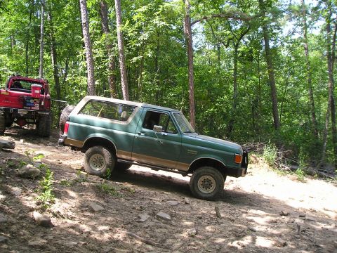 1989 Ford Bronco - A green 1989 Ford Bronco navigates a rocky, wooded trail, followed closely by a red Jeep.