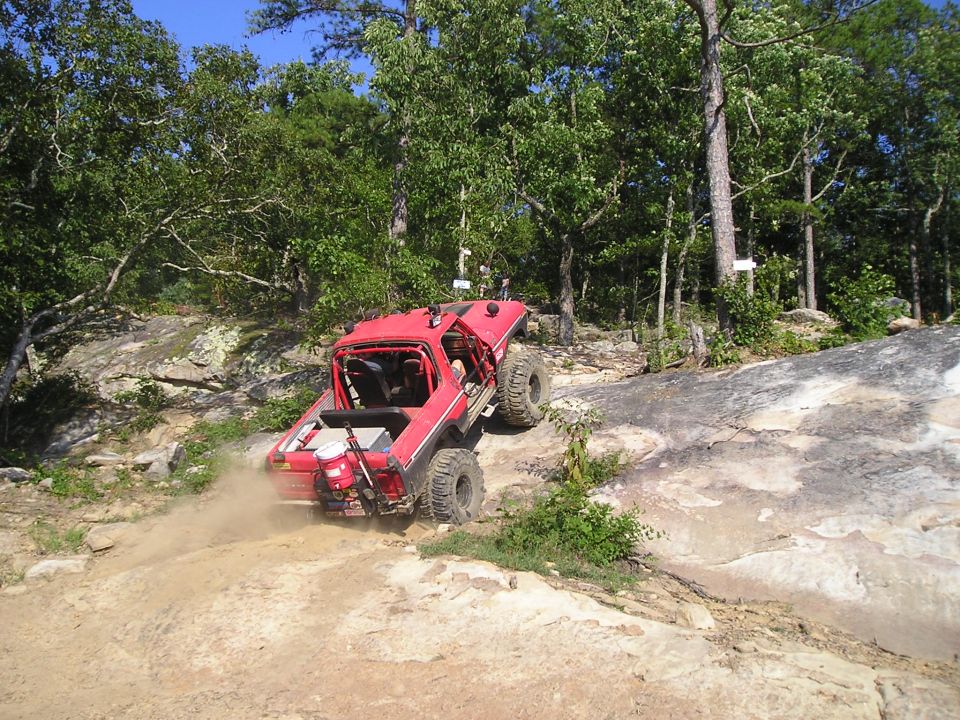 Red off-road SUV with large tires climbing a steep, rocky incline, kicking up dust