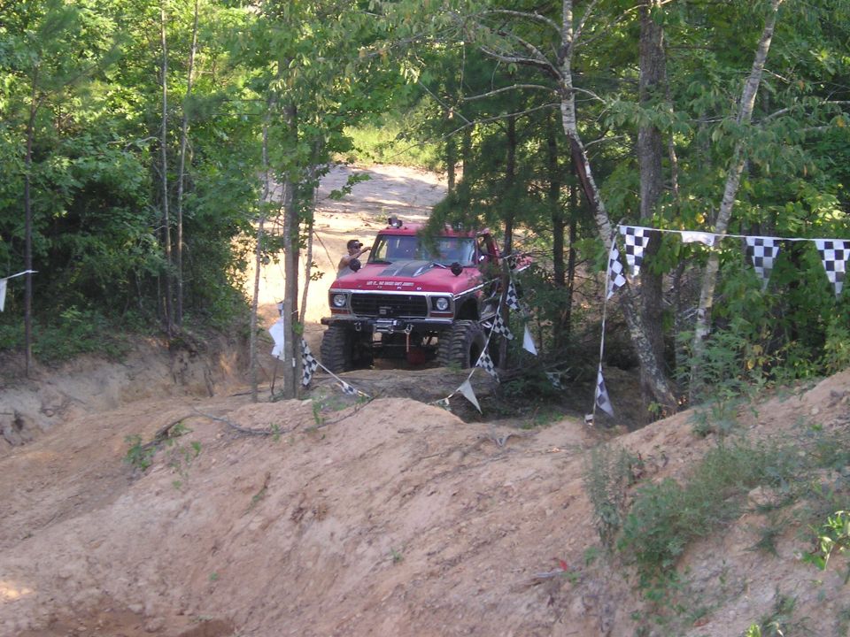 Red off-road vehicle with large tires on a dirt track, surrounded by trees. Checkered flags are visible