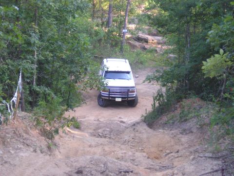 1989 Ford Bronco - A white SUV navigates a rough dirt path surrounded by dense foliage.