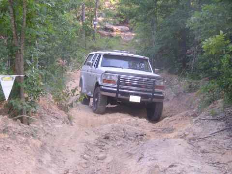 1989 Ford Bronco - A silver SUV navigates a challenging dirt path surrounded by trees.