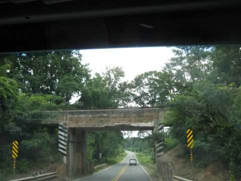 1978 Ford Bronco - A single car travels on a two-lane road beneath a concrete overpass surrounded by lush green trees.