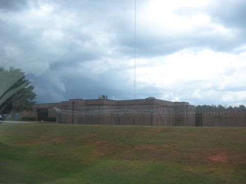 1978 Ford Bronco - A large brick building with a barbed wire fence surrounding it is visible under a dramatic, cloudy sky.