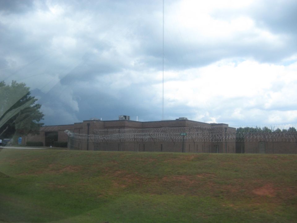 Prison building with barbed wire fence under a cloudy sky, seen from a grassy hill