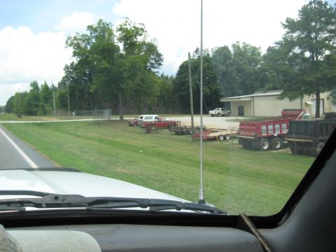 1978 Ford Bronco - Various trailers and trucks are parked in a lot beside a building and a highway.