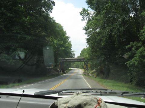 1978 Ford Bronco - View from inside a vehicle driving on a rural road towards a railway bridge, surrounded by dense green trees.