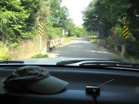 1978 Ford Bronco - View from inside a vehicle looking towards a bridge on a tree-lined road, with a hat on the dashboard.