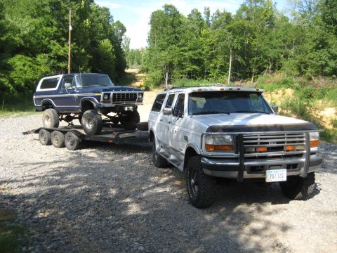 1978 Ford Bronco - A lifted blue Ford Bronco is transported on a trailer, pulled by a white Ford Excursion on a gravel road.