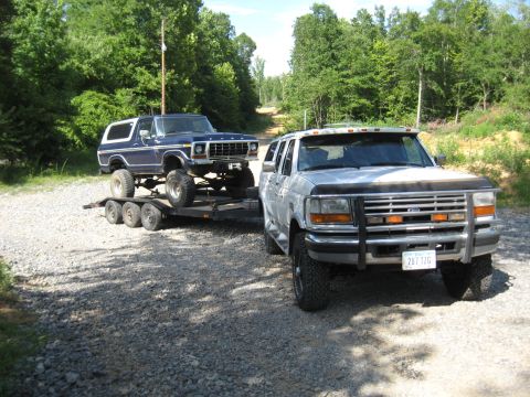 1978 Ford Bronco - A blue 1978 Ford Bronco is transported on a trailer, pulled by a silver Ford F-Series truck on a gravel road.