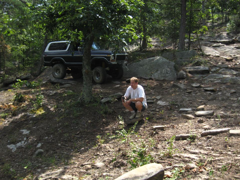 1978 Ford Bronco parked on rocky terrain under a tree, man crouching nearby